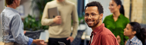 Young cheerful mixed race man holding digital tablet and smiling at camera while working with colleagues in the modern office, selective focus photo