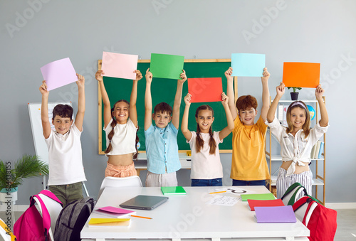 Cheerful school children standing in row in classroom and holding different colorful mockup banners. Team of happy primary junior students play games, study new words and raise text copy space posters