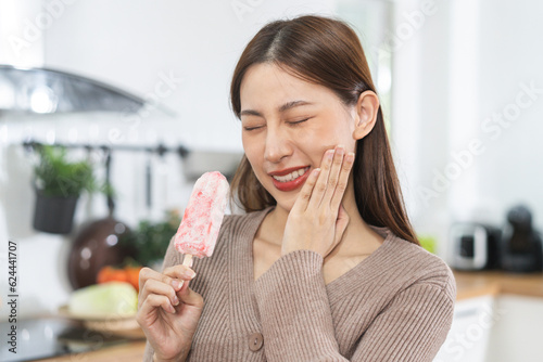 Face expression suffering from sensitive teeth and cold, asian young woman, girl hand touching her cheek, feeling hurt, pain eating ice cream, lolly. Toothache molar tooth at home, dental problem. photo