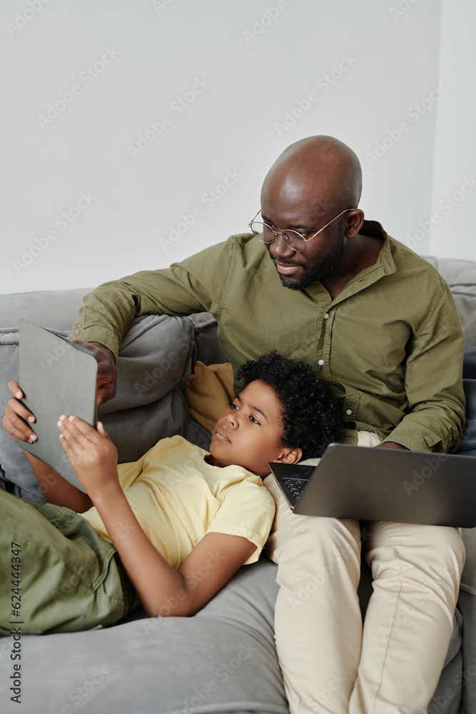 Vertical image of African American dad helping his son to use tablet pc while working on laptop on sofa in the room