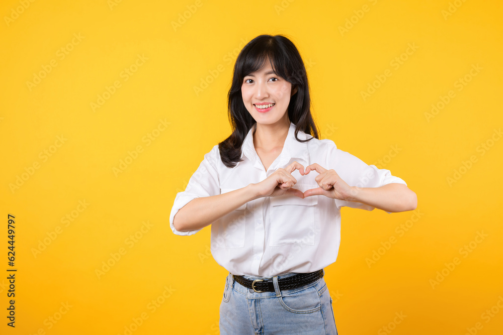 Young female healthcare. Asian woman wearing white shirt feels happy and romantic shapes heart gesture expresses tender feeling poses isolated on yellow background. People affection and care concept. Young female healthcare. Asian woman wearing white shirt feels happy and romantic shapes heart gesture expresses tender feeling poses isolated on yellow background. People affection and care concept.
