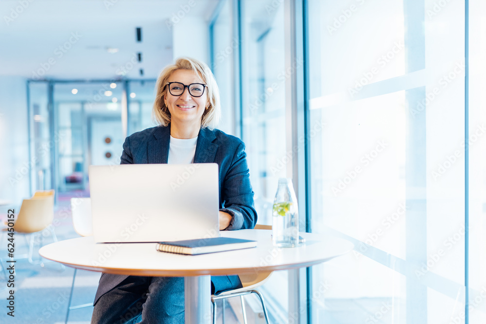© okrasiuk - Portrait of 50's confident mature businesswoman looking at camera, middle-aged experienced senior female professional working on laptop in open space office. Female entrepreneur working remotely