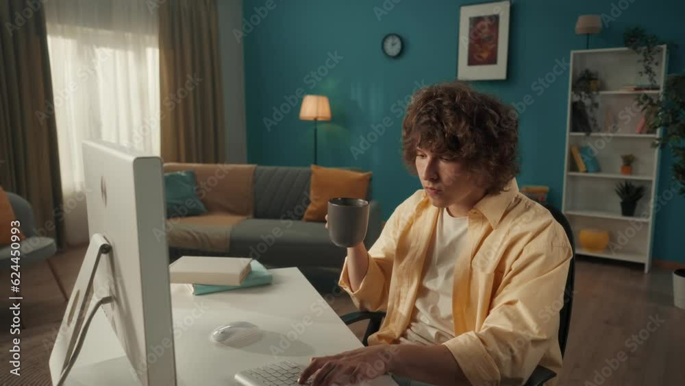 A young curly guy sits at a computer and drinks his morning coffee, tea. Man typing on keyboard, writing email, message, chatting, working online, freelancer. Portrait of a young curly man close up. A young curly guy sits at a computer and drinks his morning coffee, tea. Man typing on keyboard, writing email, message, chatting, working online, freelancer. Portrait of a young curly man close up.