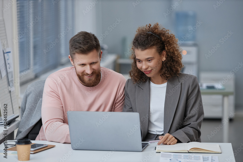Two young economists in smart casualwear looking at laptop screen while sitting by desk at working meeting and discussing online data Two young economists in smart casualwear looking at laptop screen while sitting by desk at working meeting and discussing online data