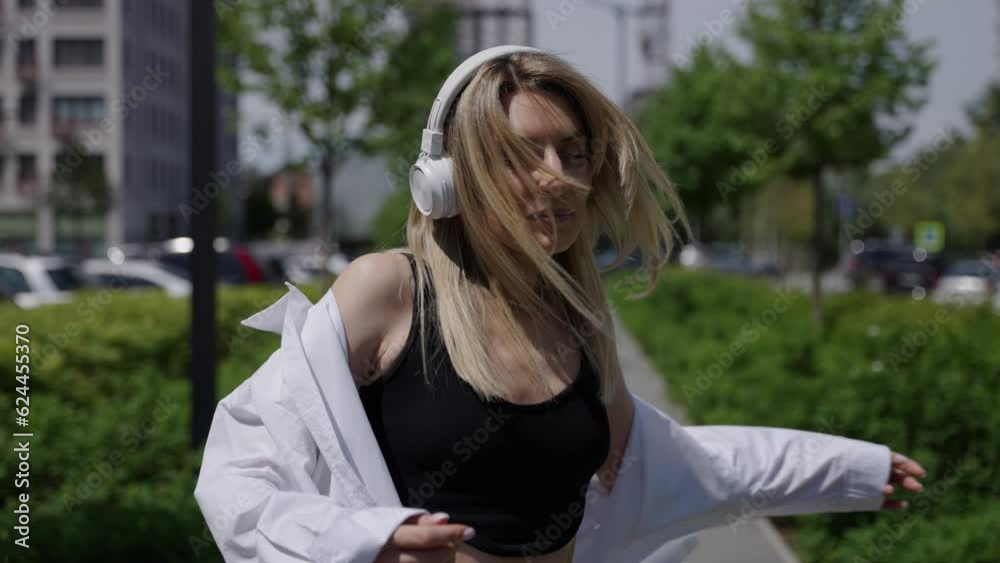 Portrait of a happy woman. Self-confidence. A girl in white headphones and blonde hair is spinning around her. Green park in the residential complex.
