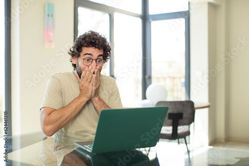 young adult bearded man with a laptop looking happy, cheerful, lucky and surprised covering mouth with both hands