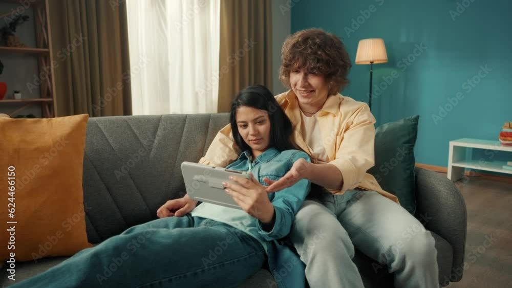 A woman and a man watch a movie, photo, video on a tablet, discuss what they saw. A young Asian woman lies on a sofa with a tablet in her hands, leaning her back on a man. Close up. A woman and a man watch a movie, photo, video on a tablet, discuss what they saw. A young Asian woman lies on a sofa with a tablet in her hands, leaning her back on a man. Close up.