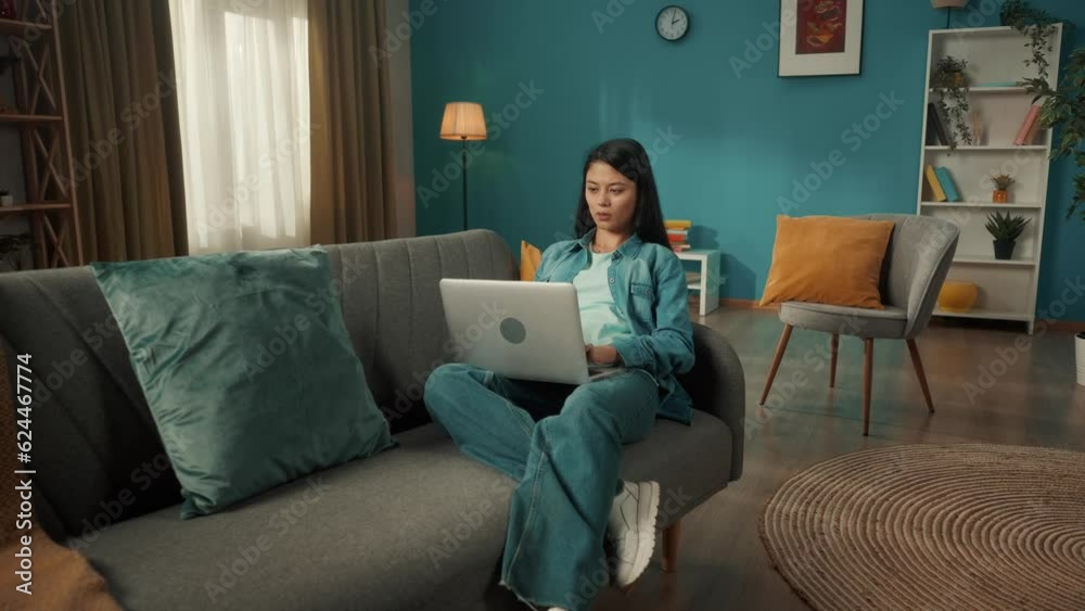 An attractive Asian woman sits on a sofa in the living room with a laptop on her lap. A woman is working, typing a text, a message, chatting. Remote work, freelancer. An attractive Asian woman sits on a sofa in the living room with a laptop on her lap. A woman is working, typing a text, a message, chatting. Remote work, freelancer.
