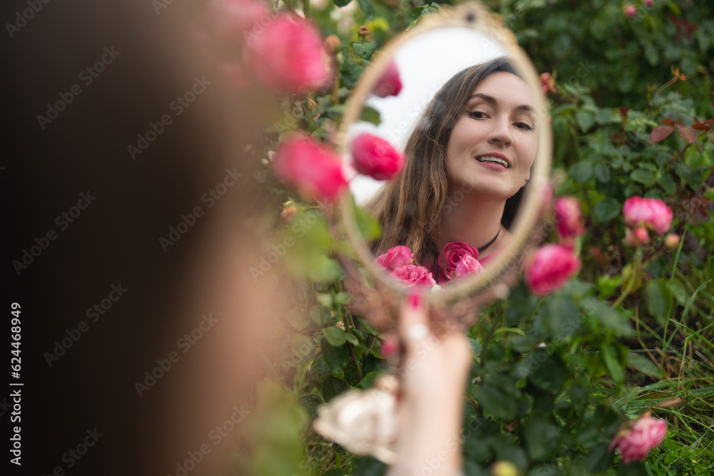 Naklejka premium a beautiful girl in a pink dress in a garden with red roses looks in the mirror