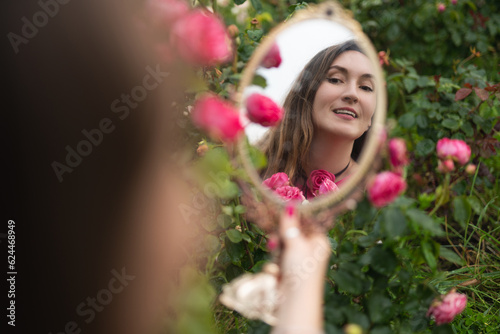 a beautiful girl in a pink dress in a garden with red roses looks in the mirror