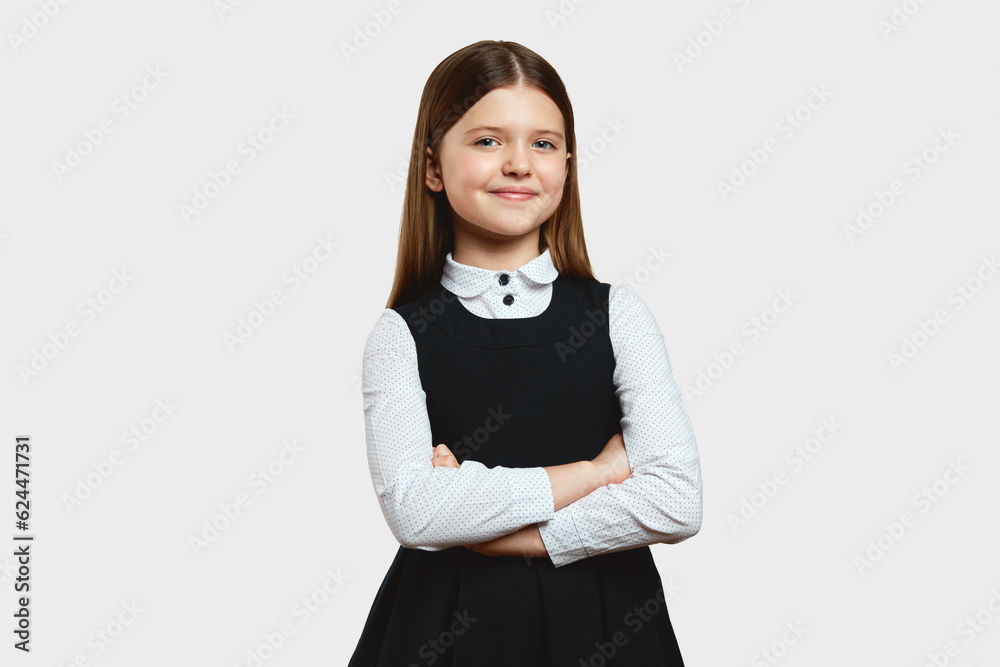 Smart focused girl in dress school uniform looking at camera while standing alone, with arms crossed against white background in studio Smart focused girl in dress school uniform looking at camera while standing alone, with arms crossed against white background in studio