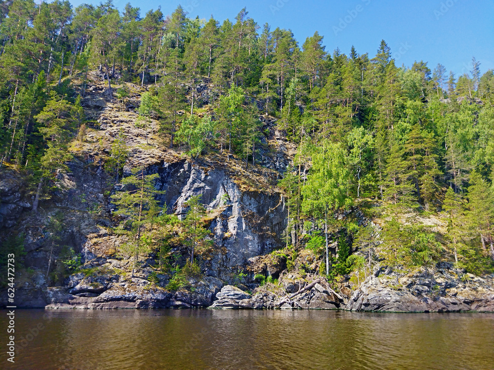 Rocky shore with large stones and trees reflected in calm water of northern lake. Beautiful summer nature of Ladoga lake. Karelia. Russia.