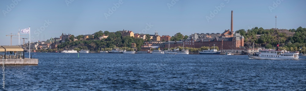 Panorama, tourist steam boat and modern ferries, industrial brick buildings, , hill Maria. district Södermalm, moored old fishing and hotel boats, a sunny summer evening in Stockholm
