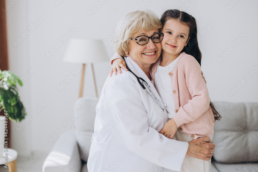 Fototapeta premium Portrait of senior nurse wearing white coat, embracing shoulders of happy little preschool patient. Smiling small girl holding favorite toy, posing for photo with attractive general practitioner.