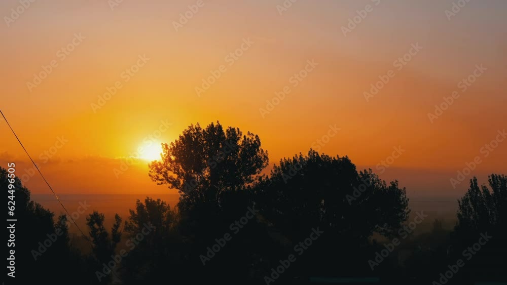Silhouette of Trees against the Backdrop of an Amazing Orange Sunrise. Time Lapse. Rays of warm yellow sun touch swaying treetops, hiding in clouds on horizon. Picturesque urban landscape. Sunset. Silhouette of Trees against the Backdrop of an Amazing Orange Sunrise. Time Lapse. Rays of warm yellow sun touch swaying treetops, hiding in clouds on horizon. Picturesque urban landscape. Sunset.