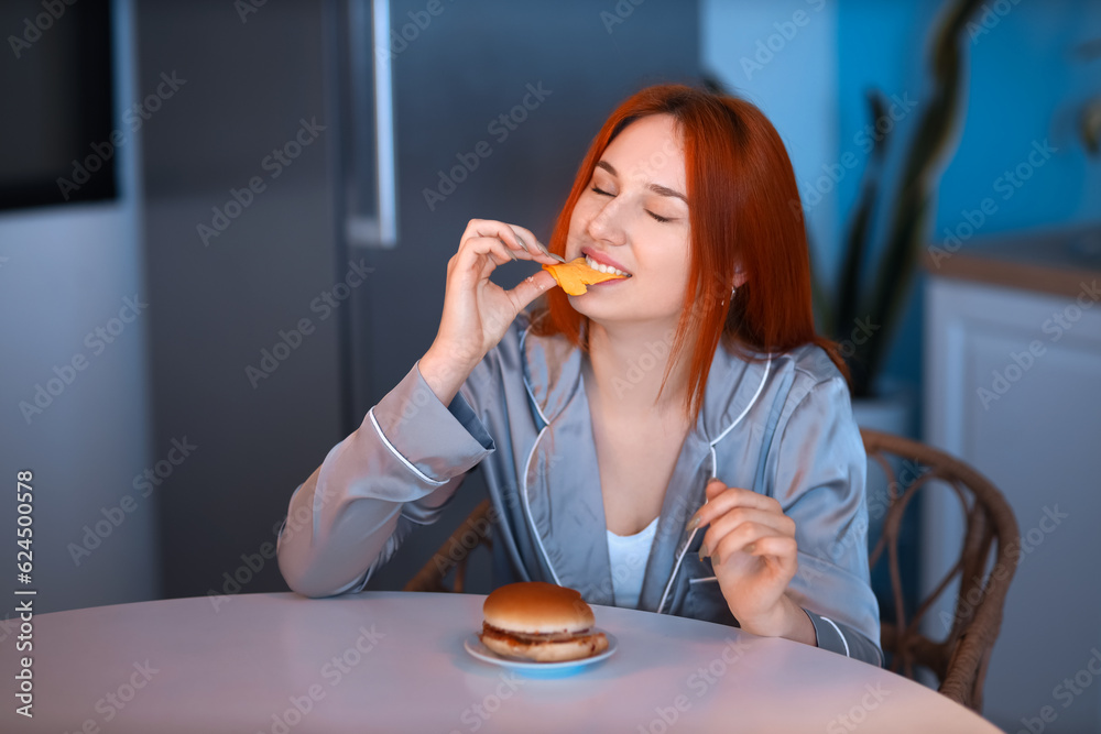 Young woman eating burger in kitchen at night Young woman eating burger in kitchen at night