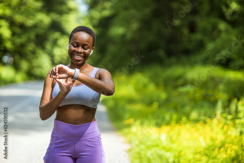 Fototapeta Naklejka Na Ścianę i Meble -  Young athlete listening to music during workout at park and adjusting smart watch. Young African-American woman setting smartwatch before jogging in summer.