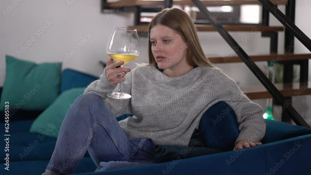 Sad beautiful woman with white wine in glass sitting on sofa thinking. Portrait of depressed young Caucasian lady alone on weekend at home indoors Sad beautiful woman with white wine in glass sitting on sofa thinking. Portrait of depressed young Caucasian lady alone on weekend at home indoors