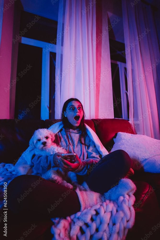 Naklejka premium Beautiful young girl at home in a dark room playing with her dog and sits in front of a monitor or TV.