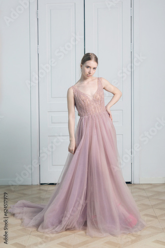 A blonde girl poses against the backdrop of white doors in a long dusty pink floor-length ball gown