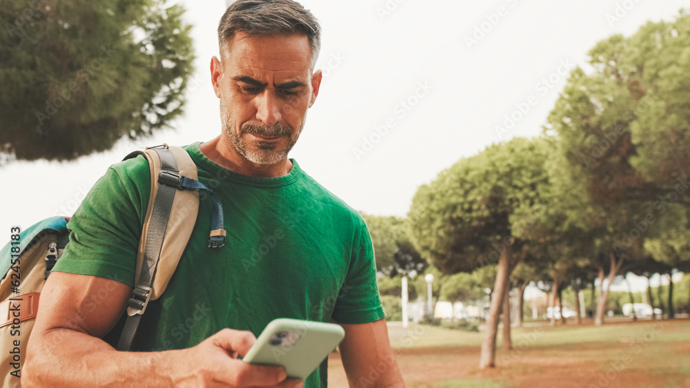 Fototapeta premium Mature man wearing casual clothes, with backpack on his shoulder, stands in the park, sends SMS from mobile phone, always online
