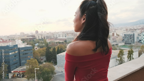 Close up, young woman standing on the balcony looking at the city at sunset, back view