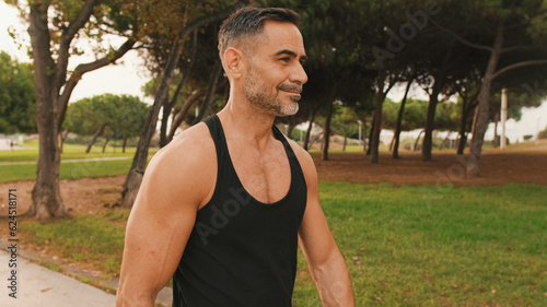 Happy mature muscular man in sportswear walking in the park after training, looking around