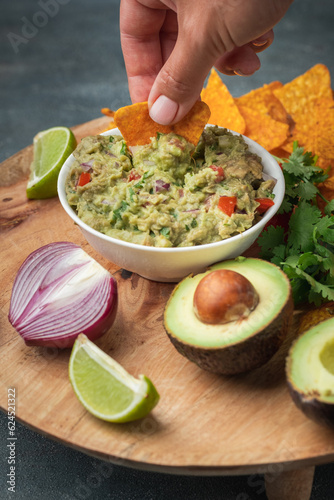 Woman hand dip nachos into tasty guacamole sauce on the wooden background. Tortilla chips next to guacamole ingredients: avocado, cilantro, onion, hot pepper and lime. Traditional Mexican food