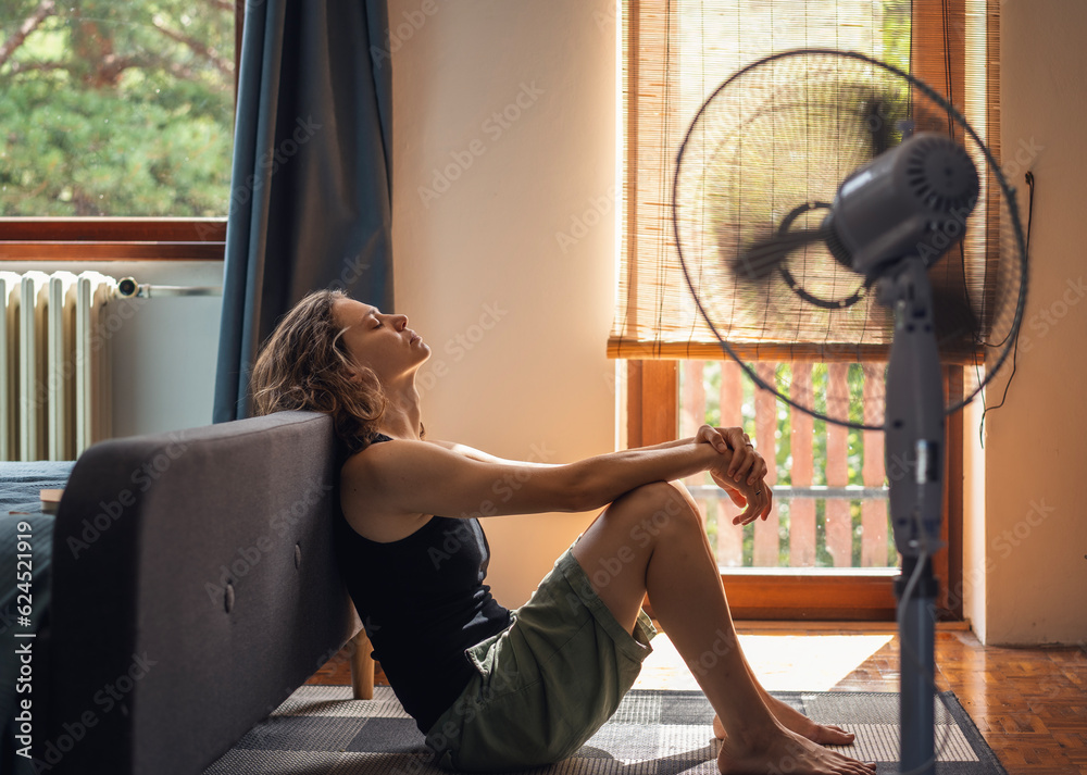 Young woman sitting at home on the floor in front of a fan saving from heat