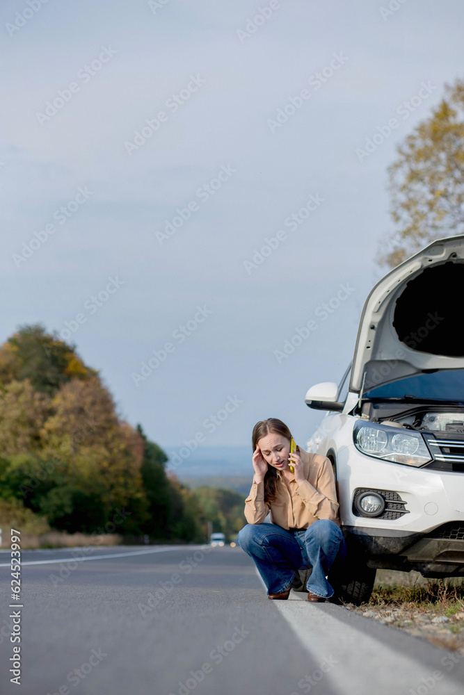 Upset young woman driver talking angrily on cell phone with assistance service near a broken car with open hood while inspecting engine having trouble with her vehicle.