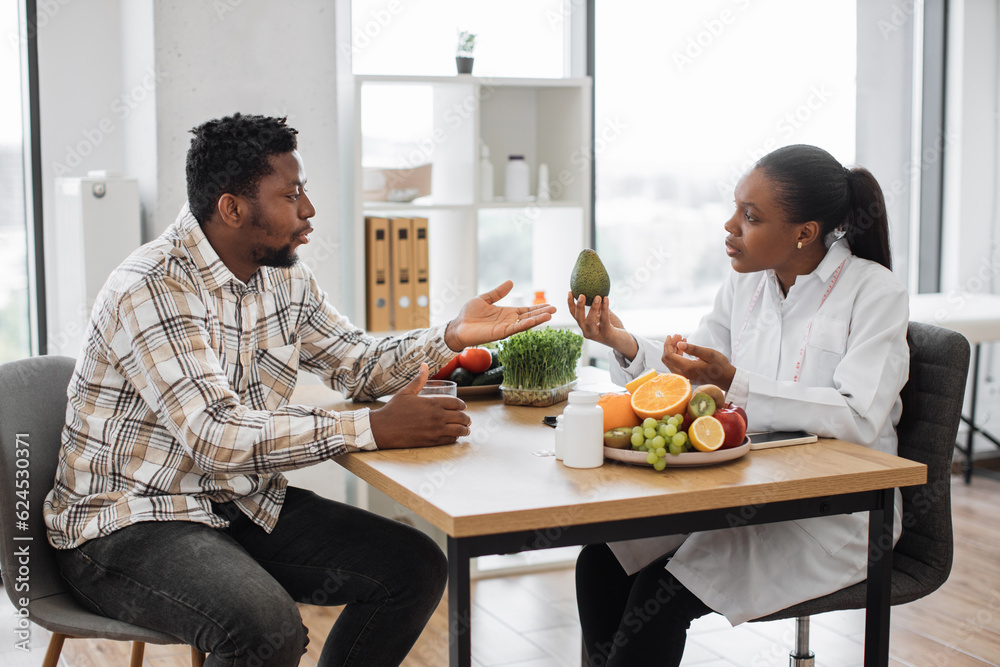 Handsome multiracial man in casual clothes visiting female dietitian in doctor's office of medical center. Curious patient asking about avocado usage for special dietary requirements on consultation. Handsome multiracial man in casual clothes visiting female dietitian in doctor's office of medical center. Curious patient asking about avocado usage for special dietary requirements on consultation.