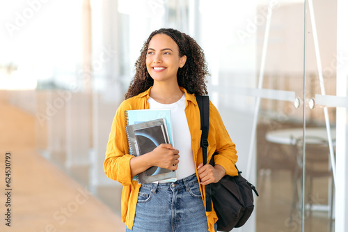 Woman university student. Happy lovely brazilian or hispanic female student, with a backpack, hold books and notebooks in her hand, stand near the university campus, looks and smile to the side