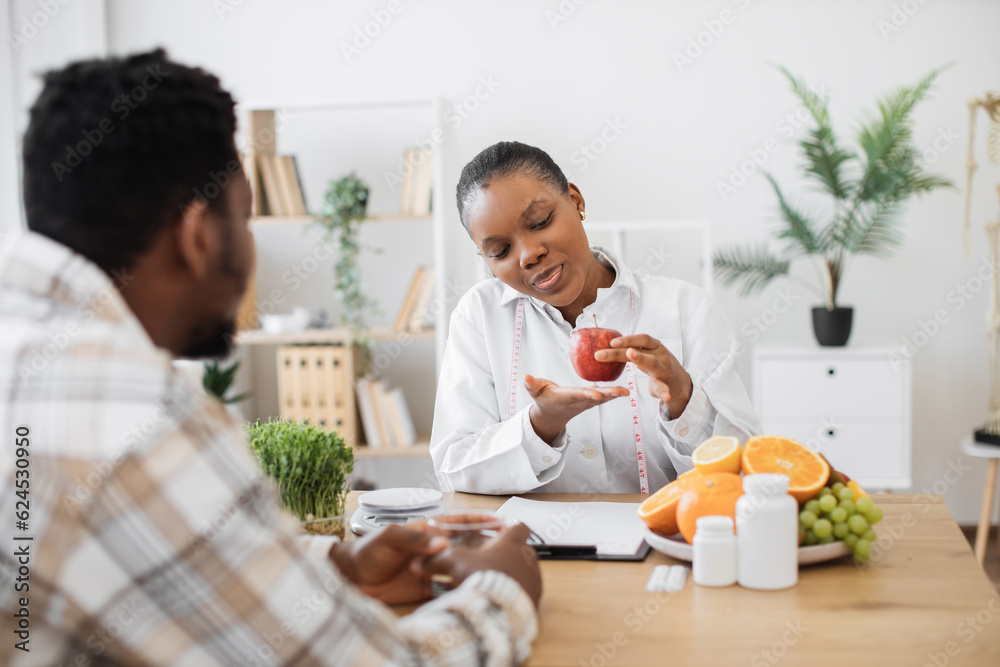 Female nutritionist holding apple with measuring tape while sitting at office desk in front of adult patient. Attractive multiracial woman in white coat promoting healthy eating with slimming foods. Female nutritionist holding apple with measuring tape while sitting at office desk in front of adult patient. Attractive multiracial woman in white coat promoting healthy eating with slimming foods.