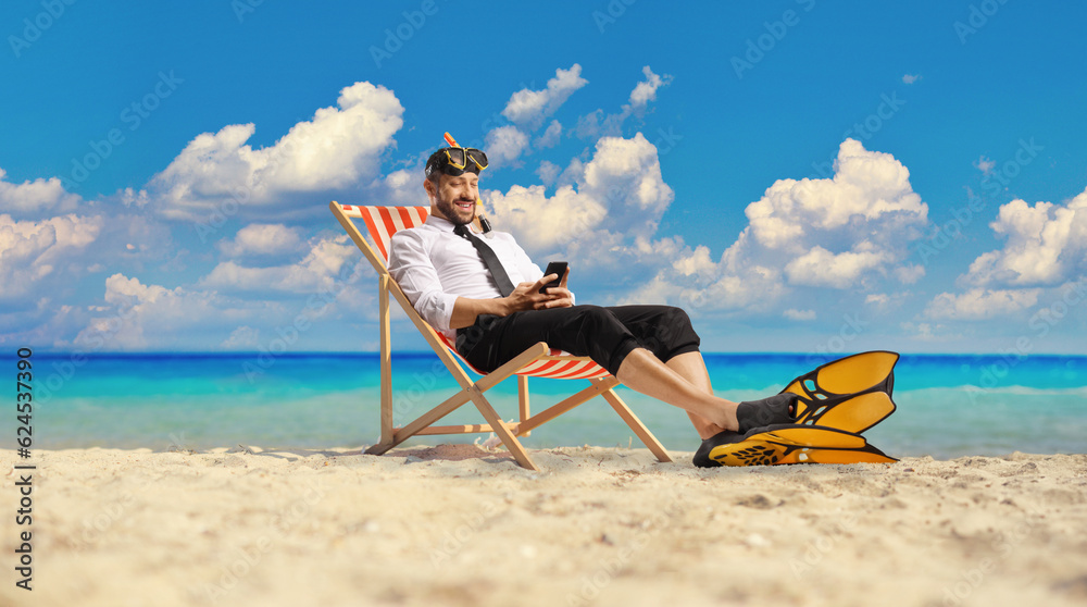 Businessman with snorkelling fins and mask sitting on a bech chair by and typing on a smartphone