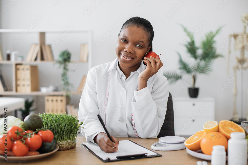 Portrait of smiling female posing with apple close to cheek while doing paperwork in doctor's office. African american nutritionist including client's preferences about fruits into meal plan. Portrait of smiling female posing with apple close to cheek while doing paperwork in doctor's office. African american nutritionist including client's preferences about fruits into meal plan.