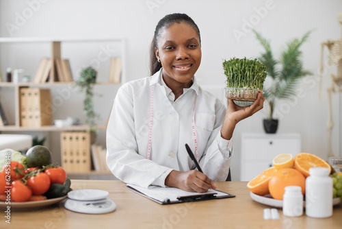 Foto Portrait of joyful female in lab coat posing with green sprouts of herbs in hand while using writing surface at desk