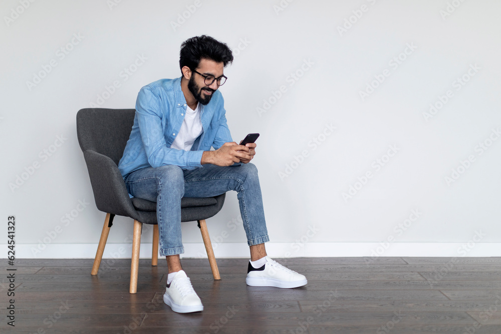 Cheerful Young Indian Man Using Cellphone While Sitting In Armchair At Home Cheerful Young Indian Man Using Cellphone While Sitting In Armchair At Home