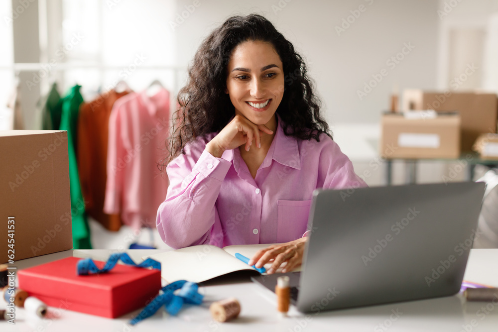 Cheerful Designer Lady Sitting At Laptop Computer In Clothes Store Cheerful Designer Lady Sitting At Laptop Computer In Clothes Store