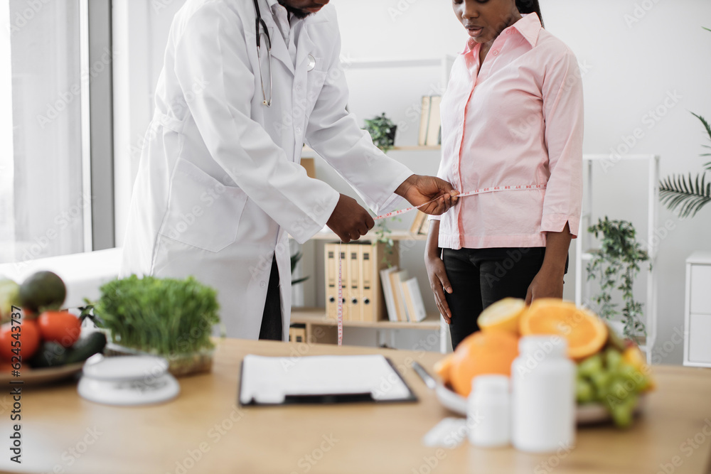 Cropped view of multicultural man in lab coat holding measuring tape on woman's waist in doctor's office. Nutrition professional giving advice patient on nutrition issues during consultation. Cropped view of multicultural man in lab coat holding measuring tape on woman's waist in doctor's office. Nutrition professional giving advice patient on nutrition issues during consultation.