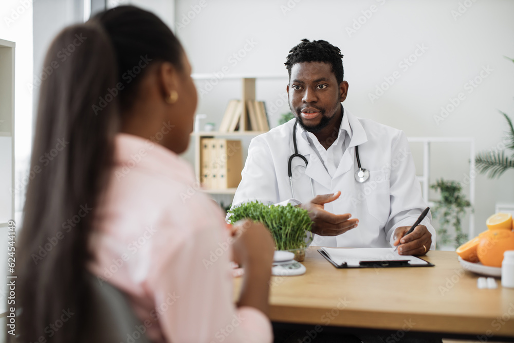 Confident man with stethoscope writing on paper clipboard during conversation with multiracial woman in office. Serious nutrition expert making notes of patient's medical history for proper meal plan. Confident man with stethoscope writing on paper clipboard during conversation with multiracial woman in office. Serious nutrition expert making notes of patient's medical history for proper meal plan.