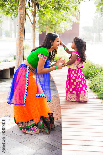 beautiful indian family mom mother with daughter girl holding hands and smiling with a bindi and traditional sari dress in front of trees and greenery