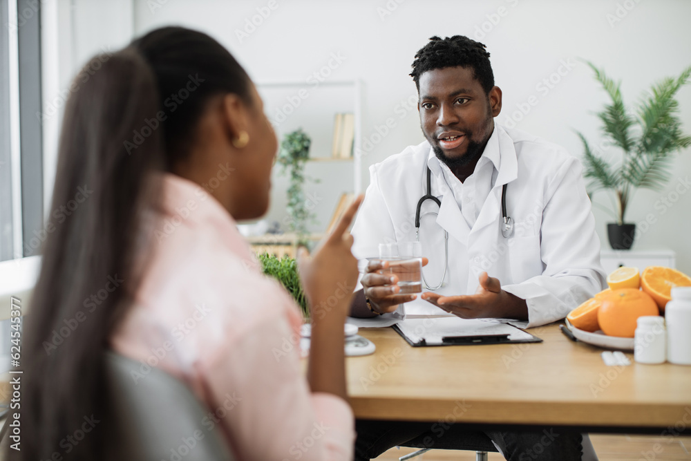 Attractive bearded man in doctor's coat gesturing at glass with fluid while meeting with multiethnic patient. Smiling nutritionist recommending clean water as help in reducing calorie intake on diet. Attractive bearded man in doctor's coat gesturing at glass with fluid while meeting with multiethnic patient. Smiling nutritionist recommending clean water as help in reducing calorie intake on diet.