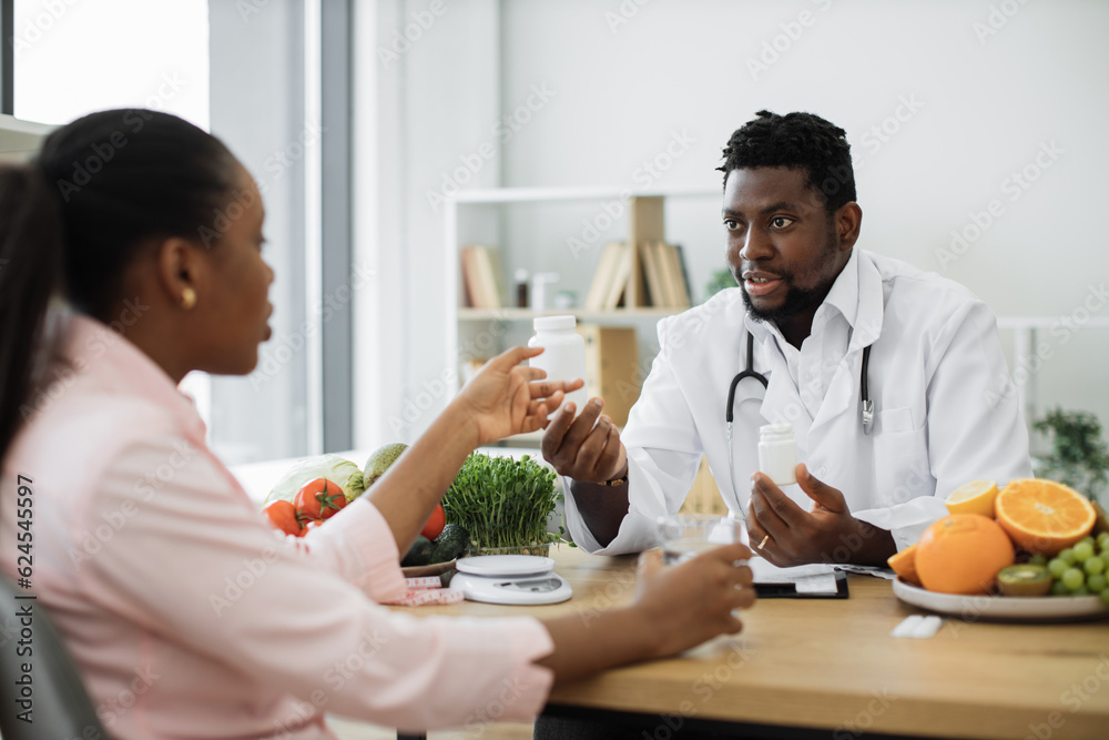 Handsome african american man in white coat giving pills bottle to charming woman in consulting room of hospital. Male nutrition professional improving overall health with dietary supplements. Handsome african american man in white coat giving pills bottle to charming woman in consulting room of hospital. Male nutrition professional improving overall health with dietary supplements.