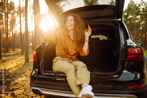 Woman is resting in trunk of car with phone. Happy tourist chatting via video call or taking a selfie in the back seat of a car. Concept of traveling by car, active lifestyle. © maxbelchenko