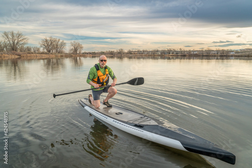 senior man (mid 60s) with a stand up paddleboard is finishing his paddling workout at a sunset, early spring lake scenery in Colorado.