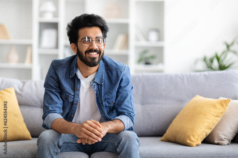 Handsome Smiling Indian Man Sitting On Couch At Home And Looking Away