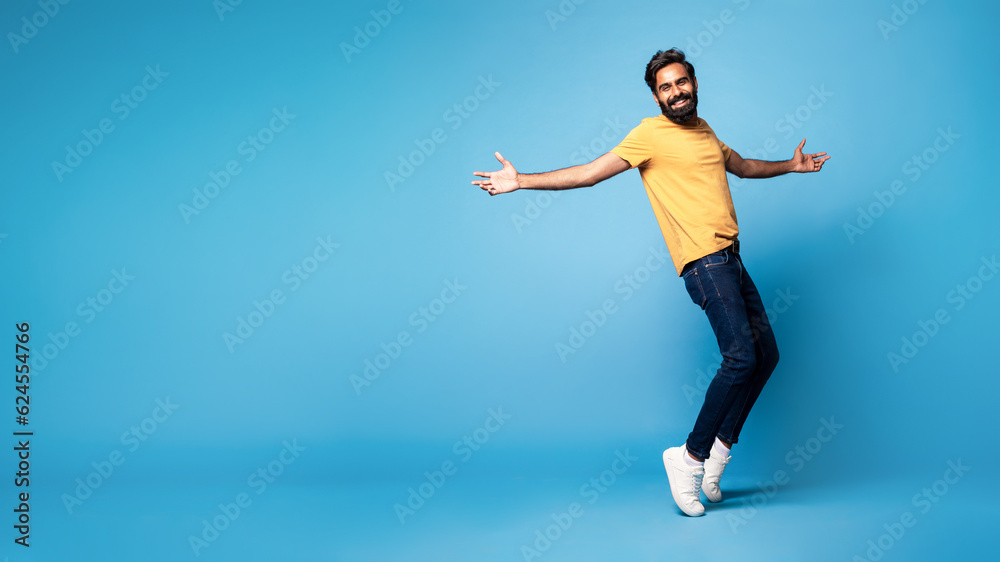 Joyful middle aged indian man in casual standing on toes and smiling at camera, panorama with free space