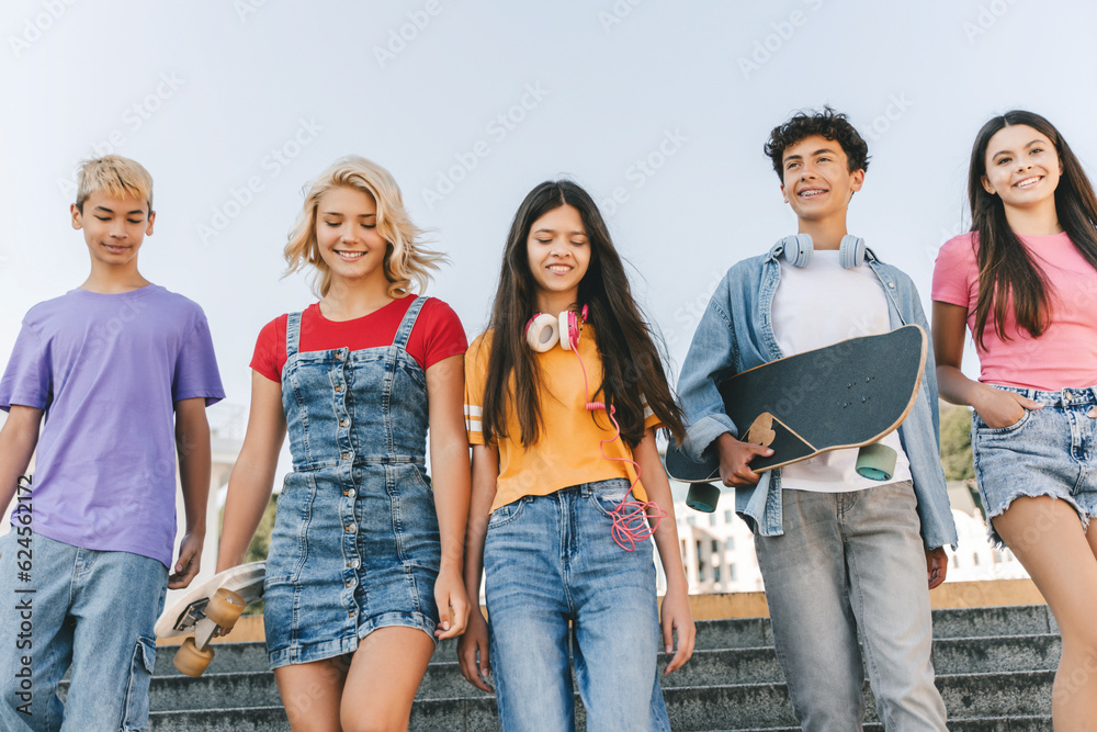 Obraz premium Group of smiling multiracial teenage friends wearing colorful t shirts talking, communication, holding skateboards walking on the street. Happy stylish boys and girls outdoors. Friendship concept