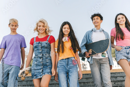 Group of smiling multiracial teenage friends wearing colorful t shirts talking, communication, holding skateboards walking on the street. Happy stylish boys and girls outdoors. Friendship concept