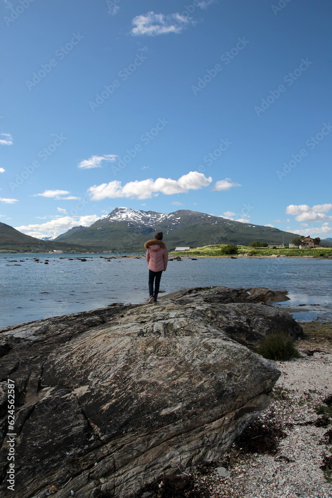 Obraz premium A girl stands on a stone on the seashore and looks at the mountains on a sunny day, Northern Norway